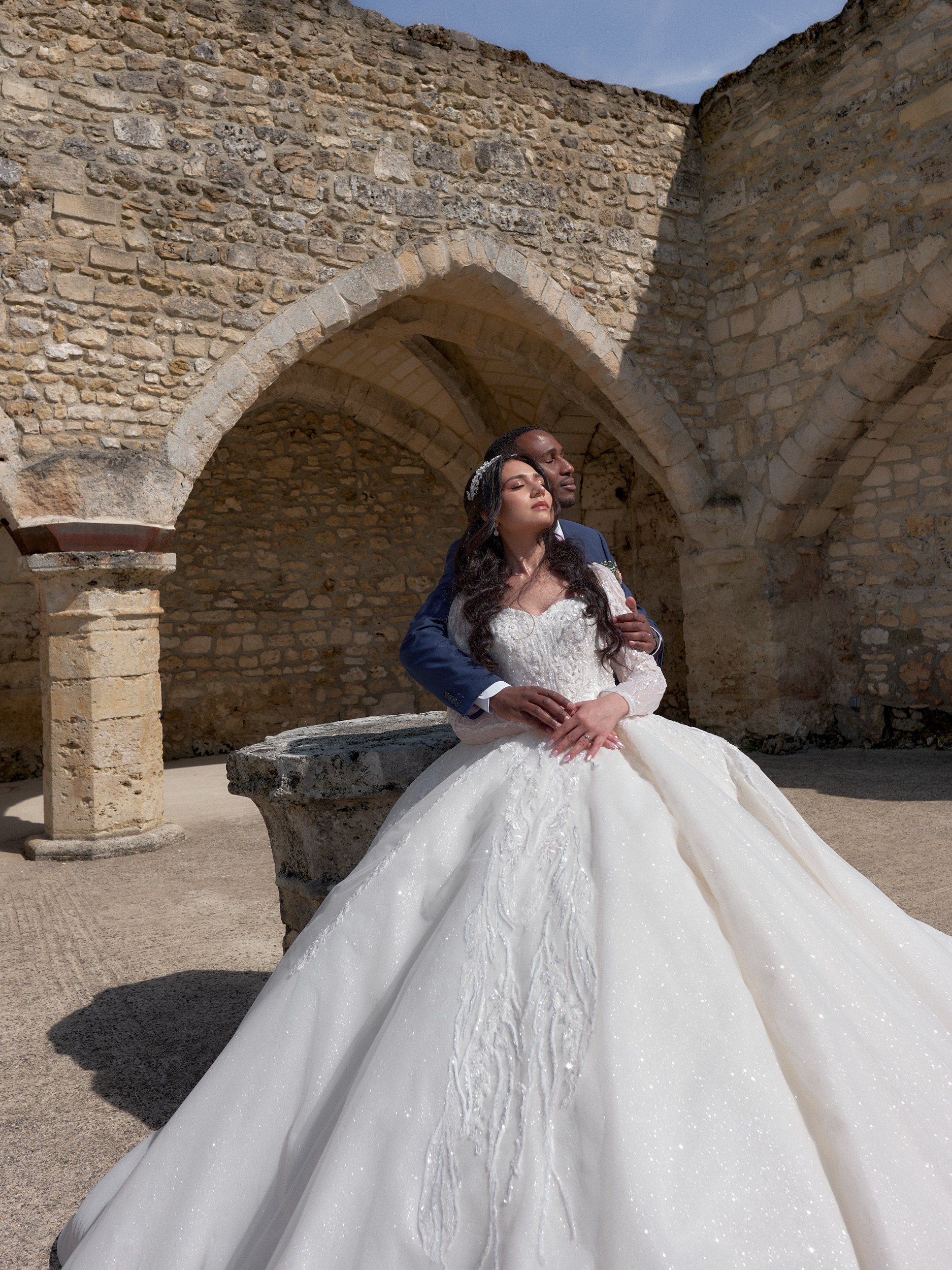 Mariés enlacés devant une arche en pierre, la mariée au premier plan.