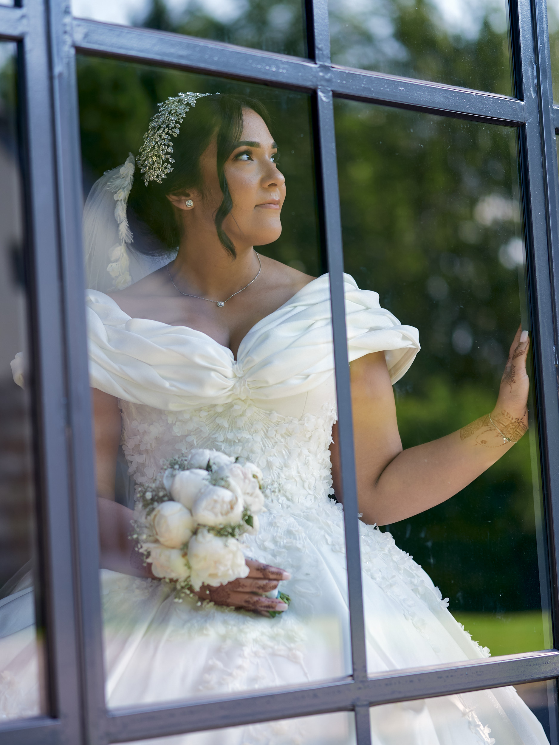 Mariée tenant un bouquet blanc, photographiée à travers les carreaux d’une fenêtre.