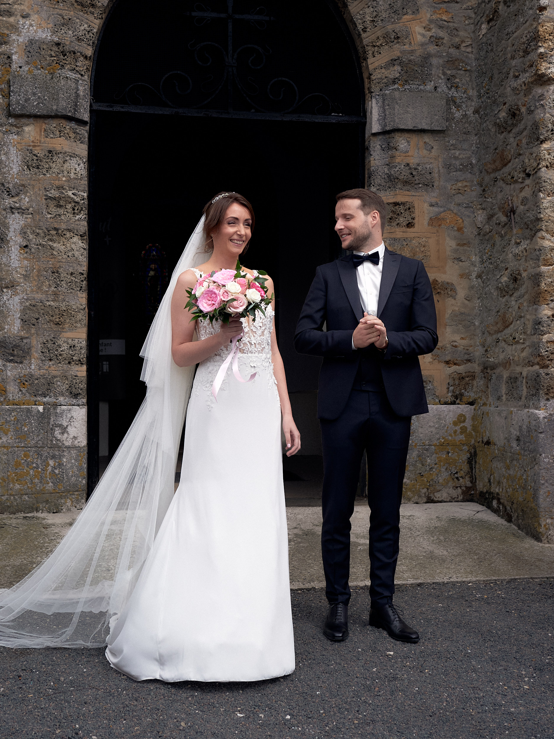 Mariés souriants devant le portail d’une église en pierre, la mariée tenant un bouquet rose.