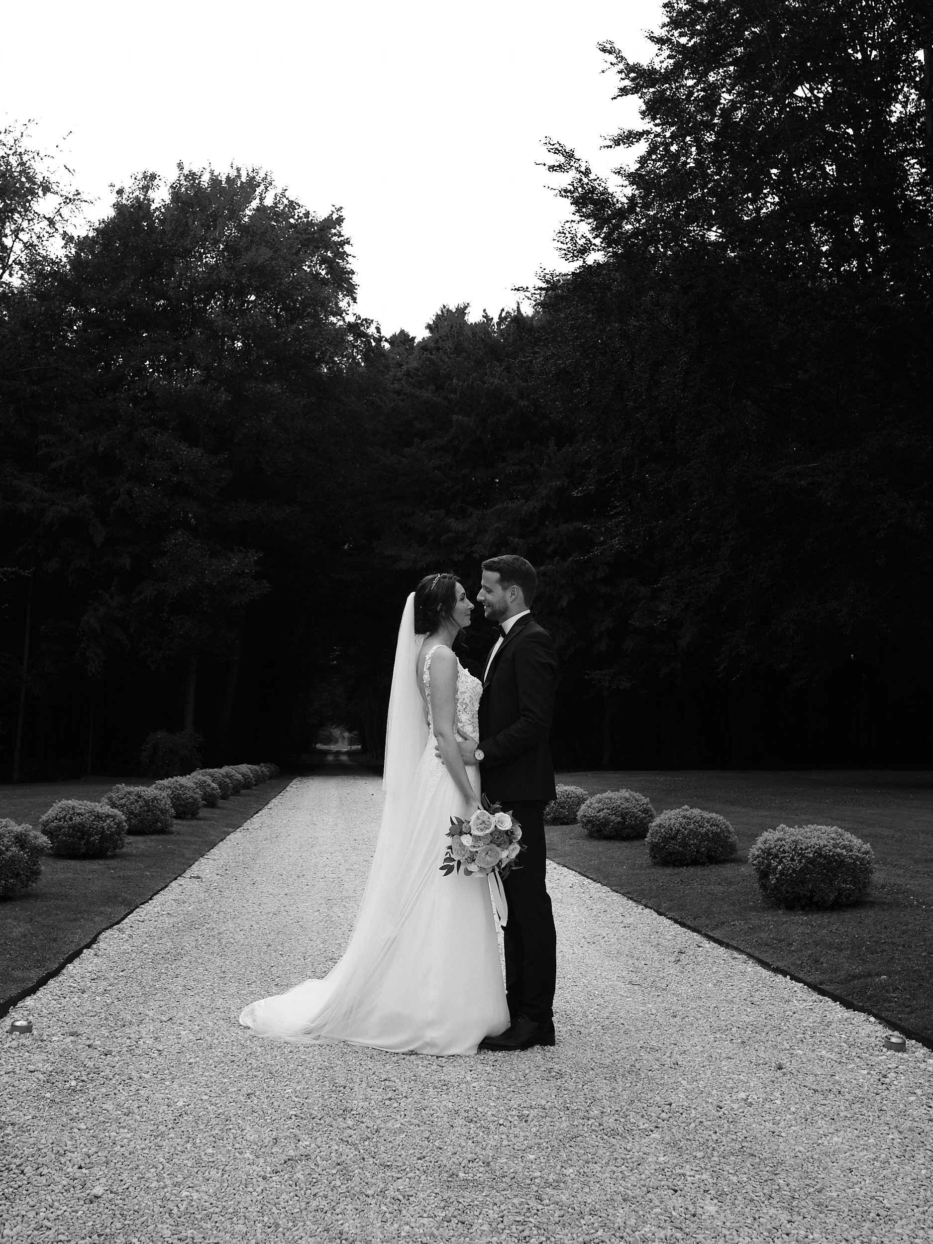 Mariés face à face sur une allée bordée d’arbres et de buissons, en noir et blanc.