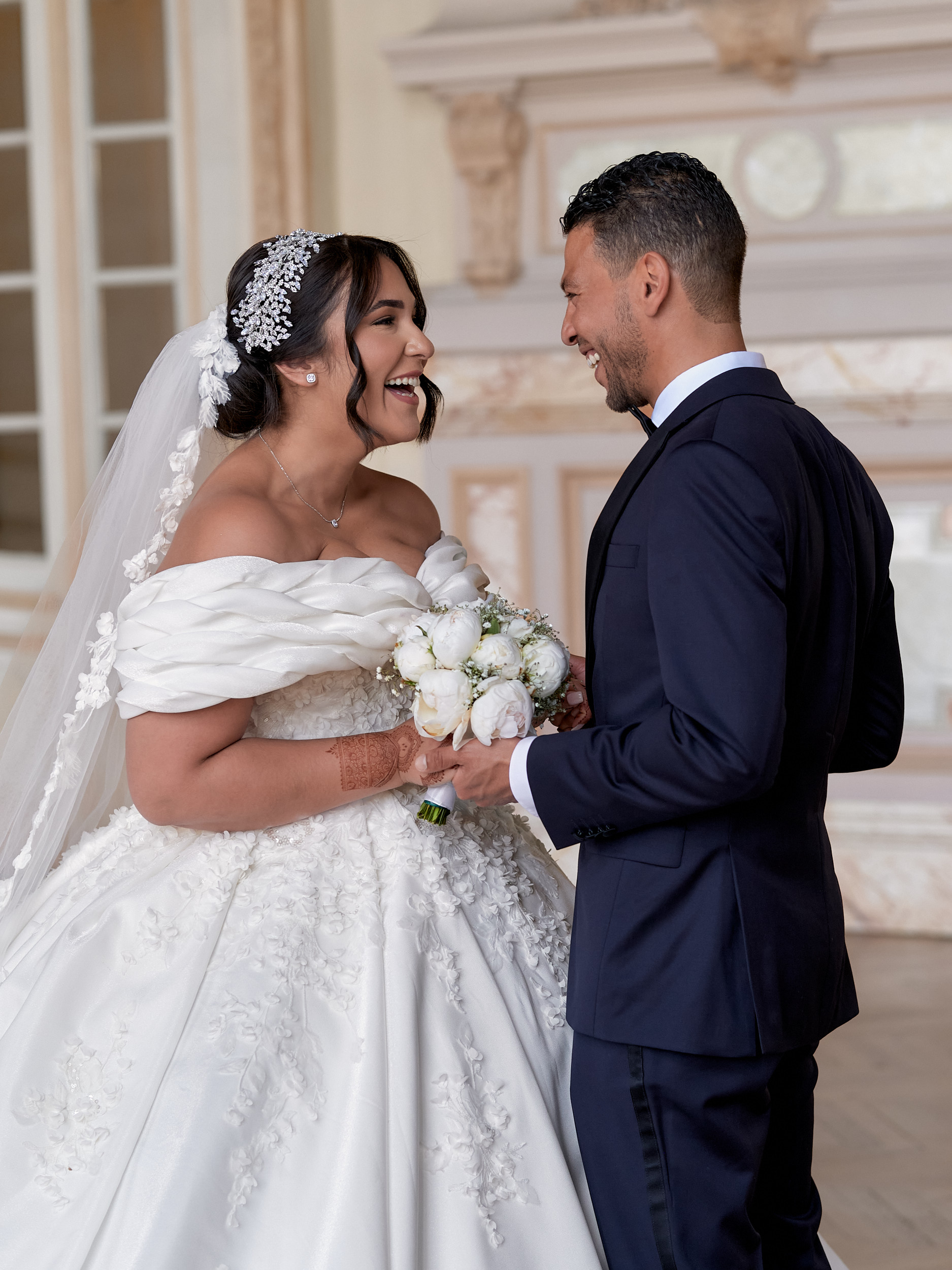 Mariés riant face à face dans un salon, un bouquet blanc entre eux.