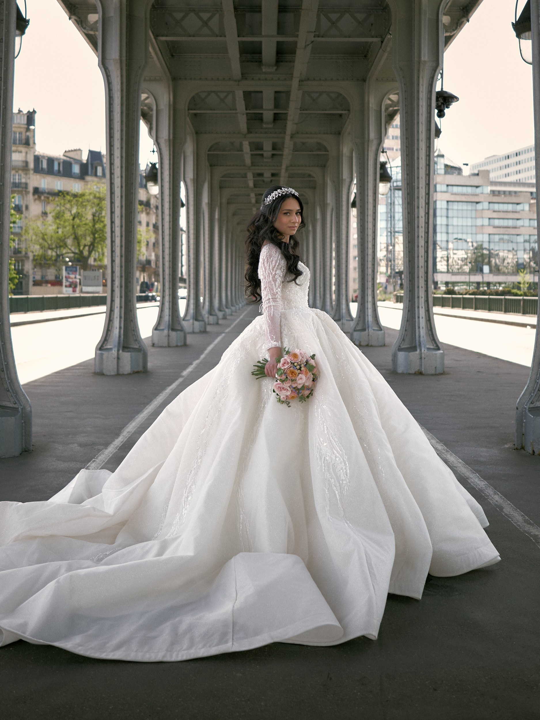Mariée tenant un bouquet sous les piliers métalliques d’un pont, sa traîne déployée.