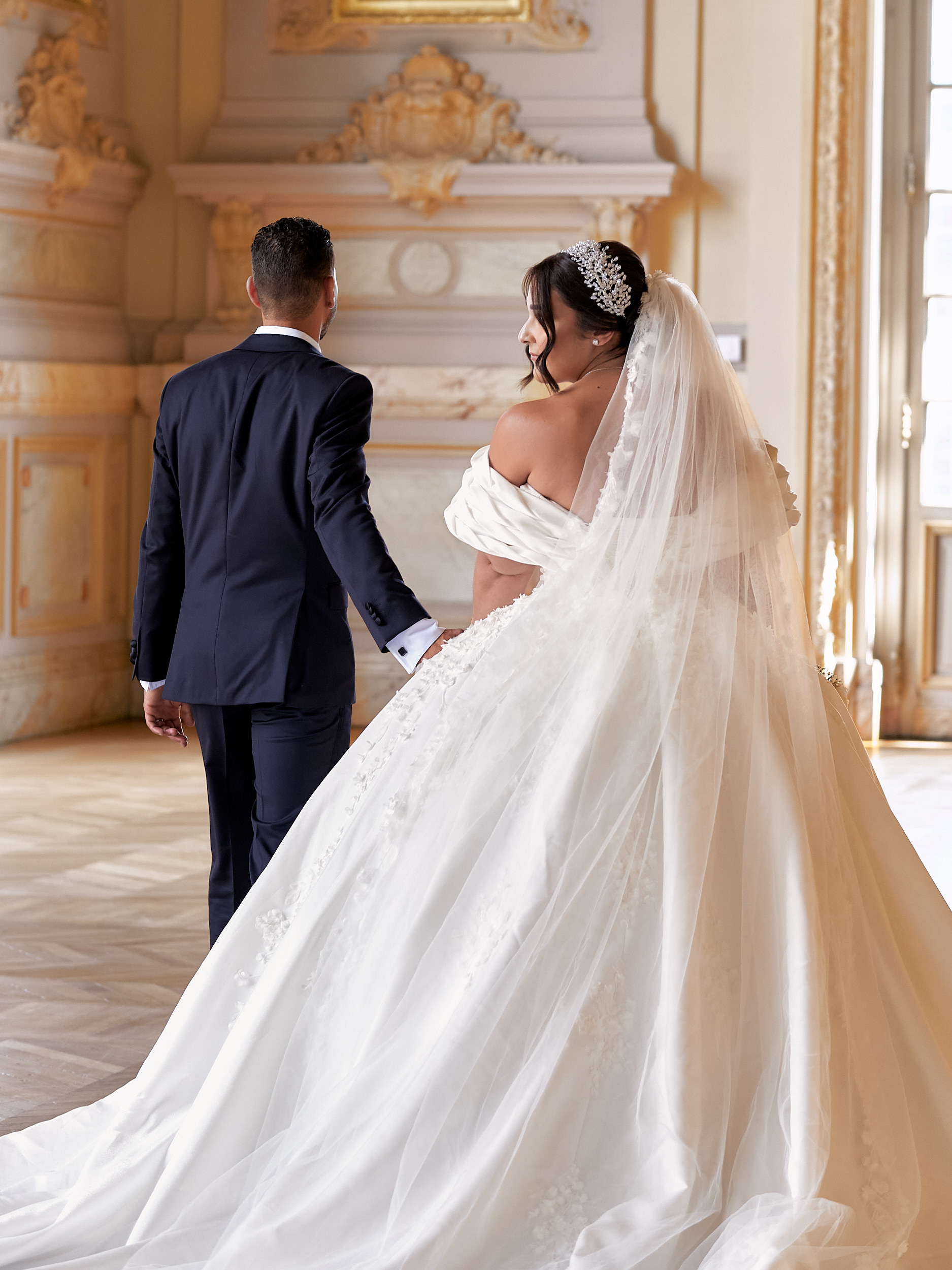 Mariés main dans la main dans un salon aux moulures dorées, la mariée se retournant.