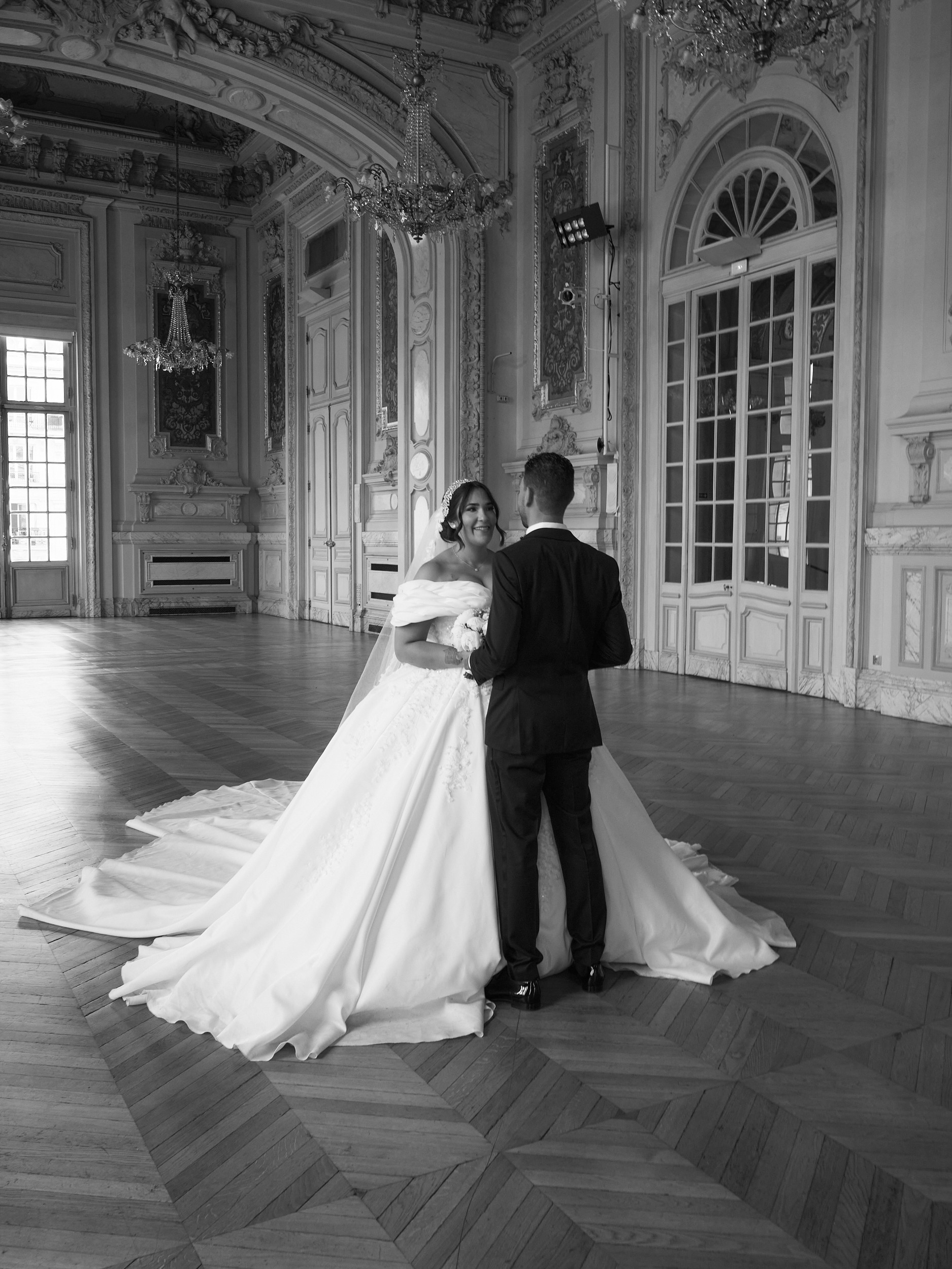 Mariés se tenant les mains dans une salle aux moulures et lustres, en noir et blanc.