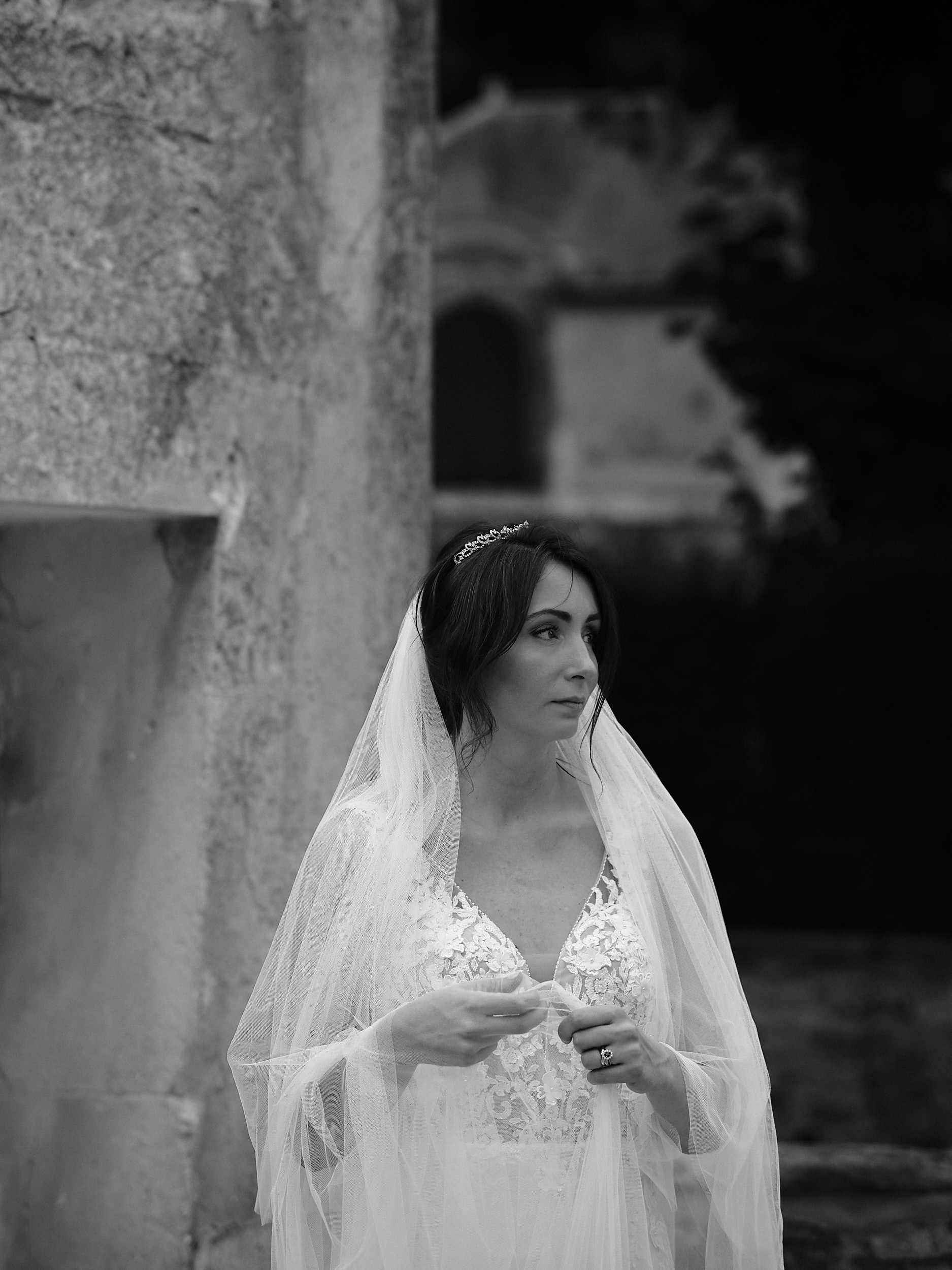 Portrait en noir et blanc d’une mariée voilée près d’un mur en pierre.