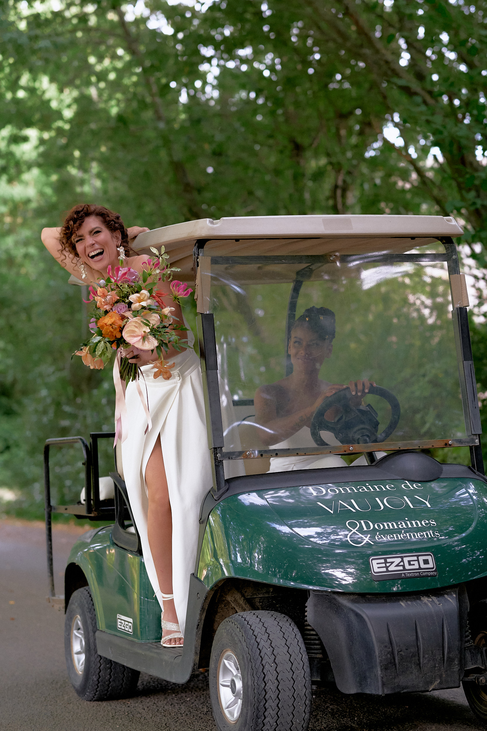Mariée souriante debout sur le marchepied d’une voiturette, un bouquet à la main.