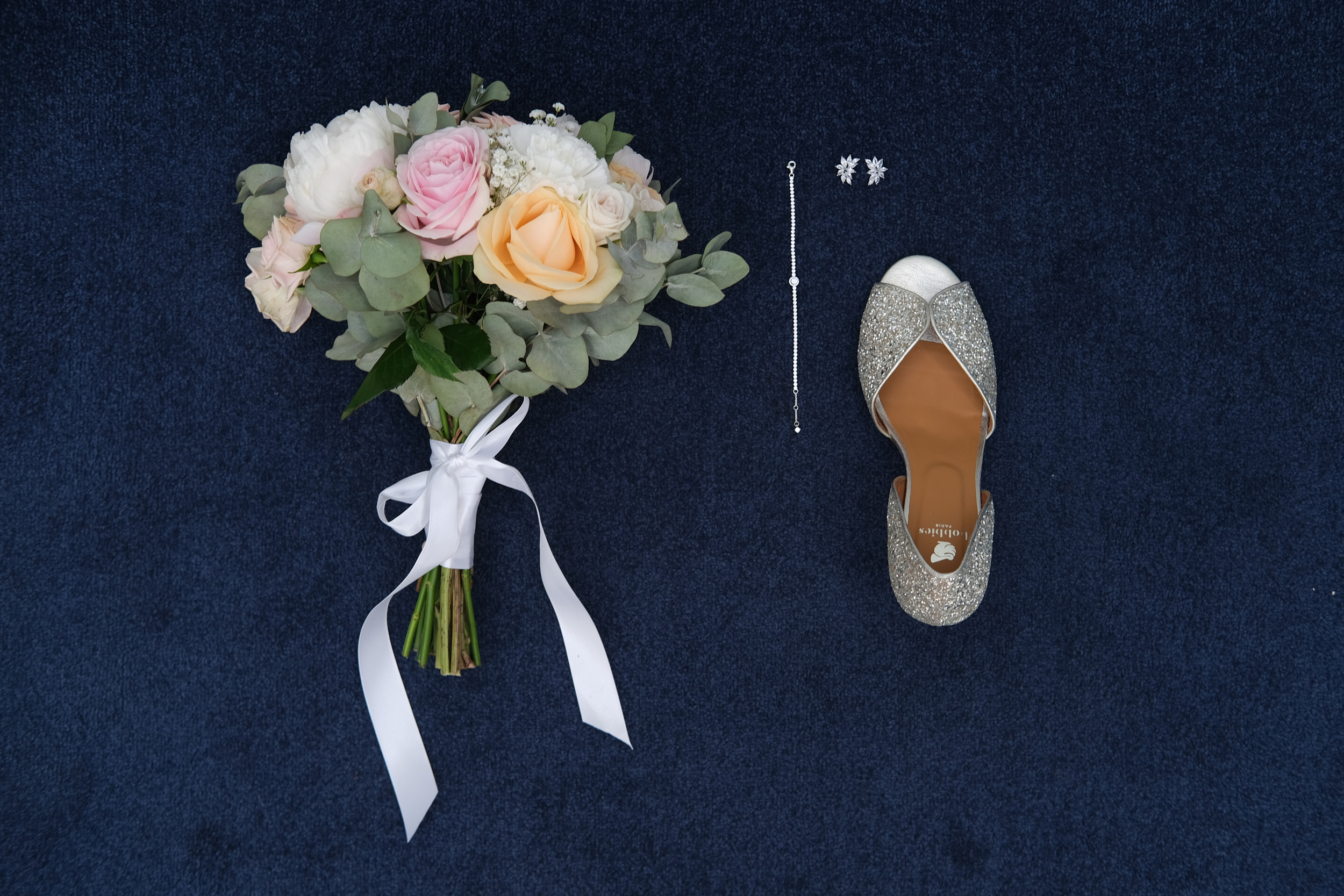 Bouquet pastel, bijoux et chaussure de mariée disposés sur un fond bleu foncé.