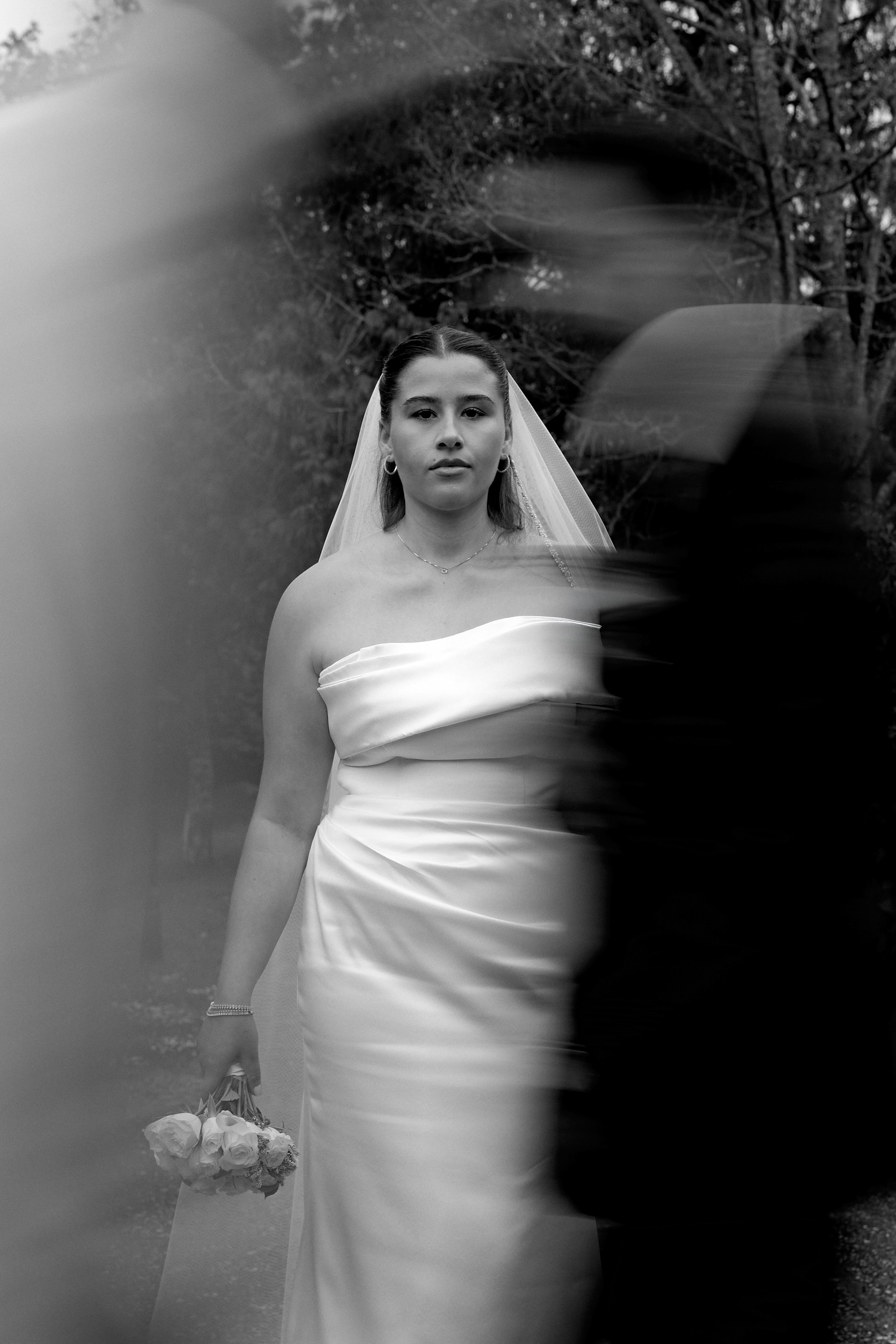 Mariée immobile tenant un bouquet, avec une silhouette floue au premier plan, en noir et blanc.