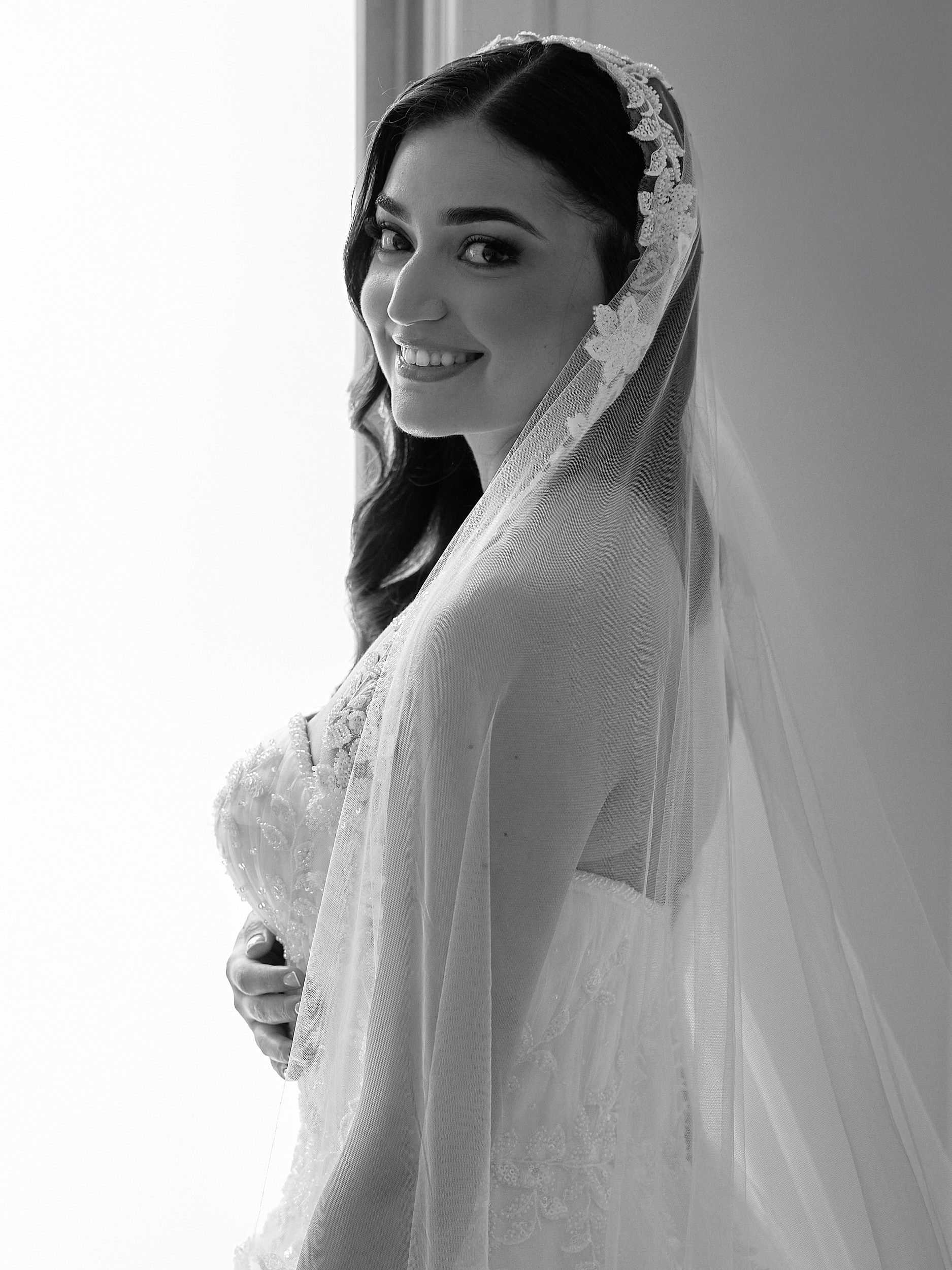 Portrait en noir et blanc d’une mariée souriante sous un voile bordé de dentelle.