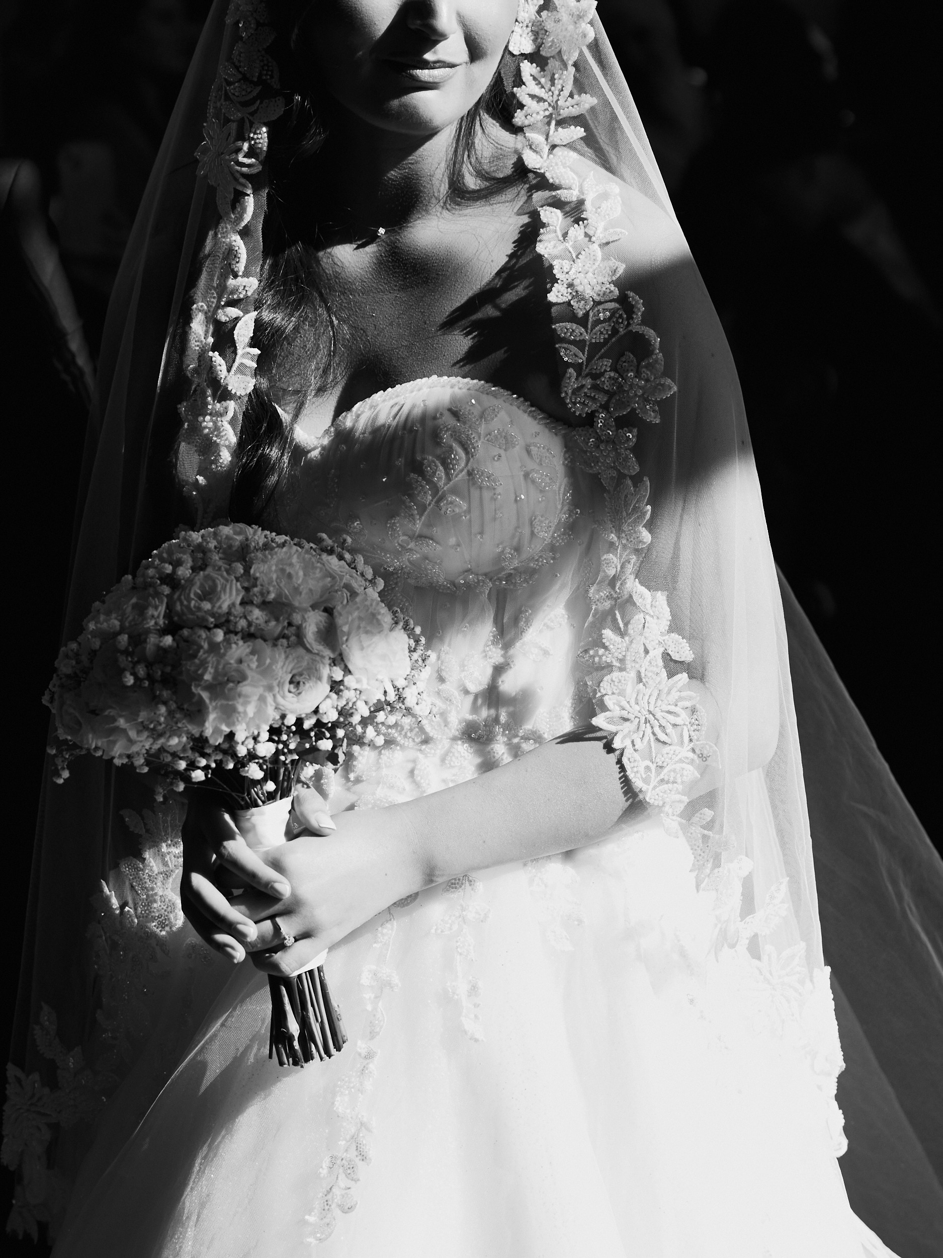 Détail en noir et blanc du bouquet, du corsage et du voile en dentelle d’une mariée, dans une lumière contrastée.