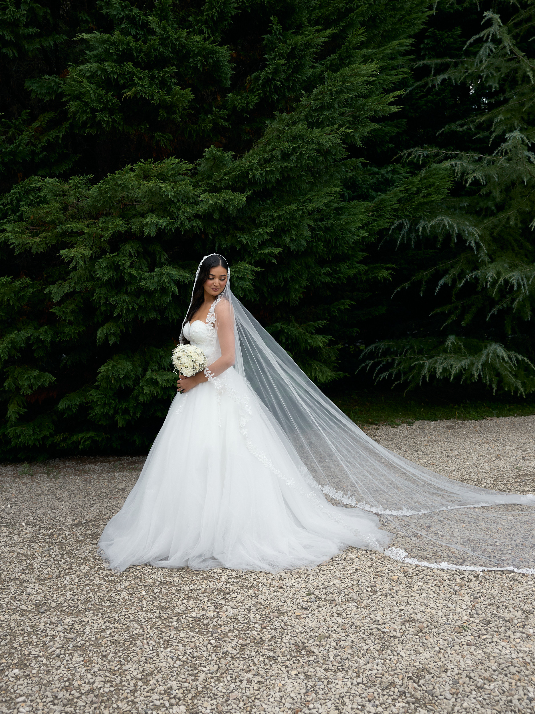 Mariée tenant son bouquet sur une allée de gravier, son long voile étendu devant les arbres.