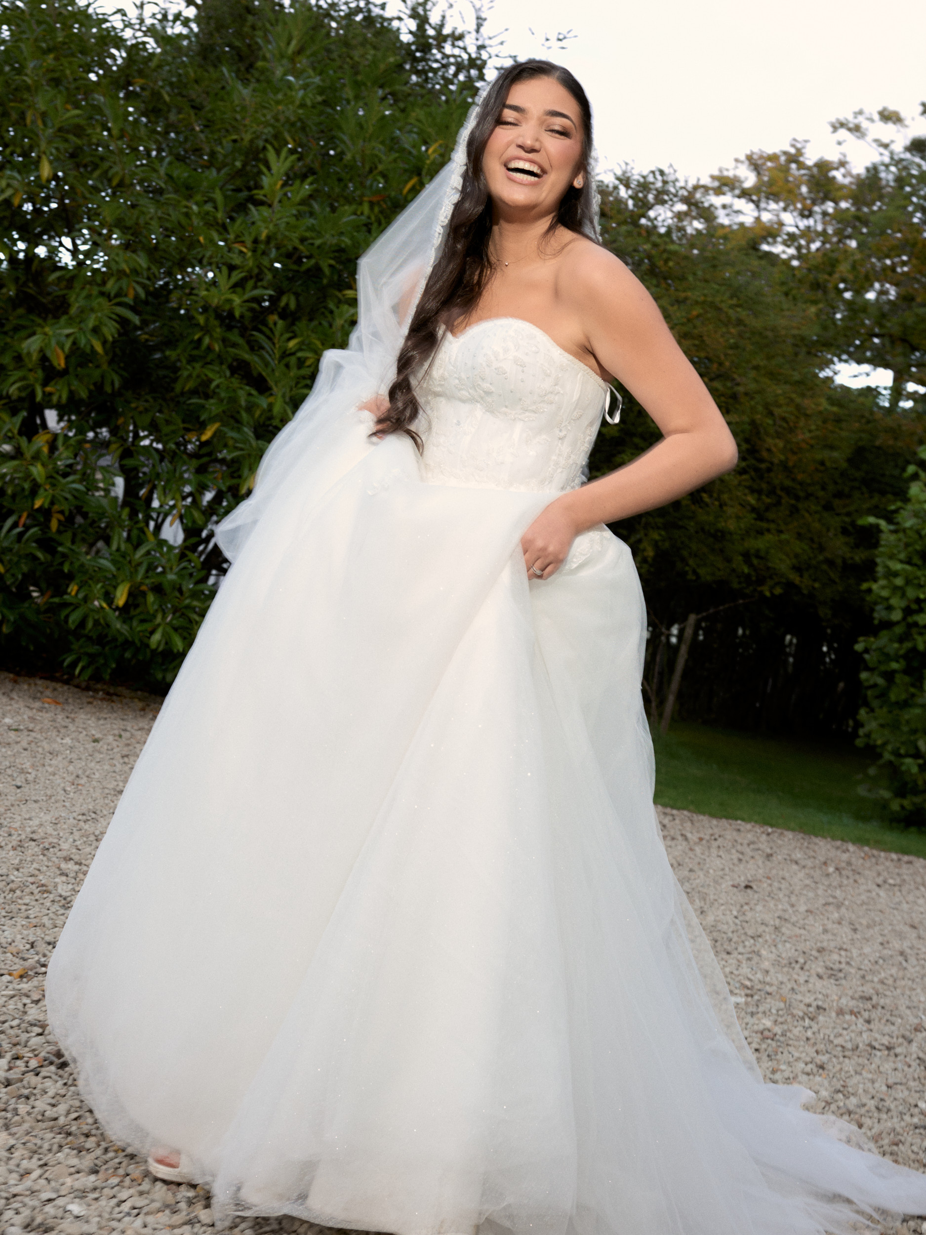Mariée riant dans un jardin, une main posée sur sa robe volumineuse.