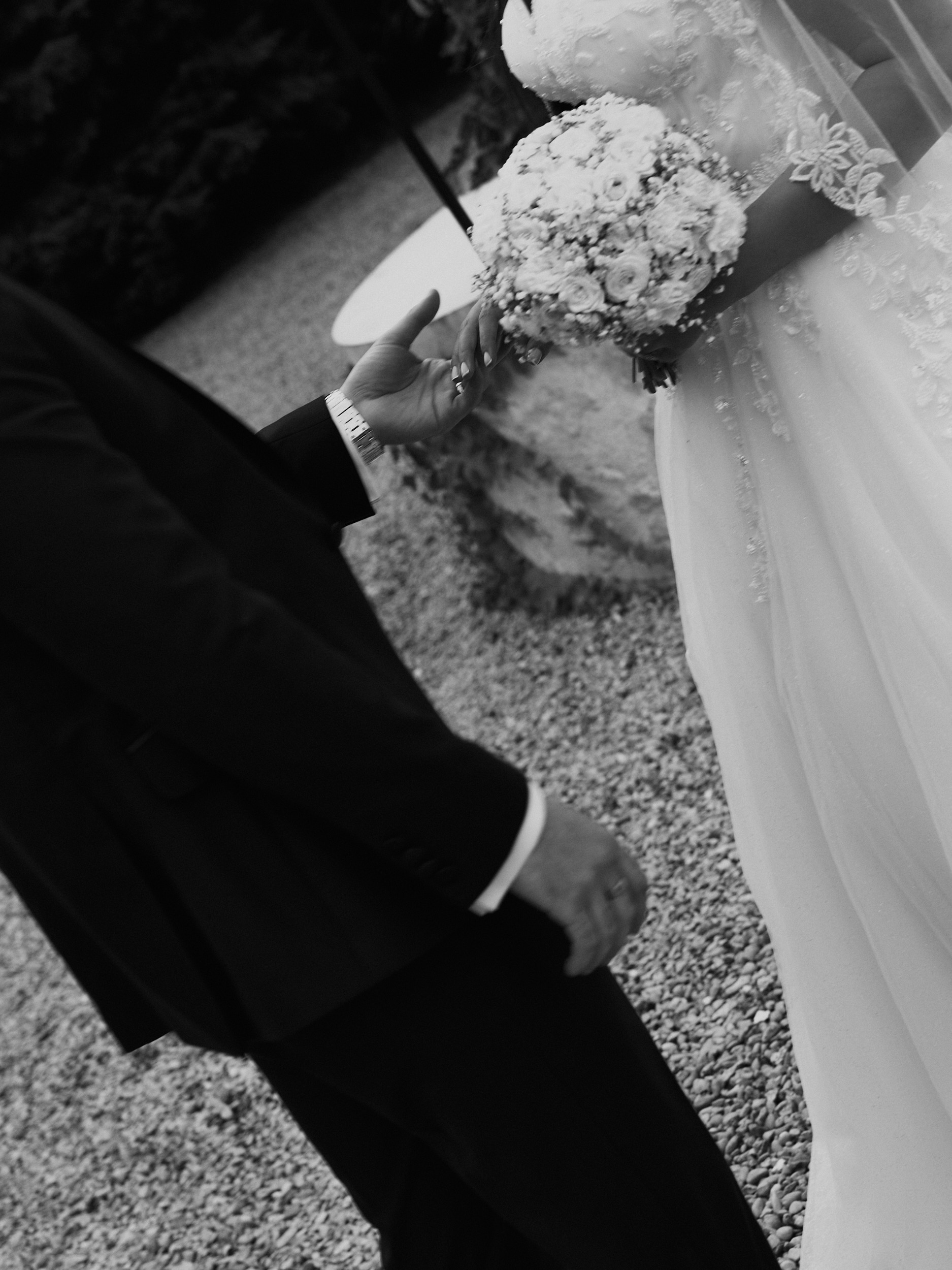 Main du marié tendue vers le bouquet de la mariée, en noir et blanc.